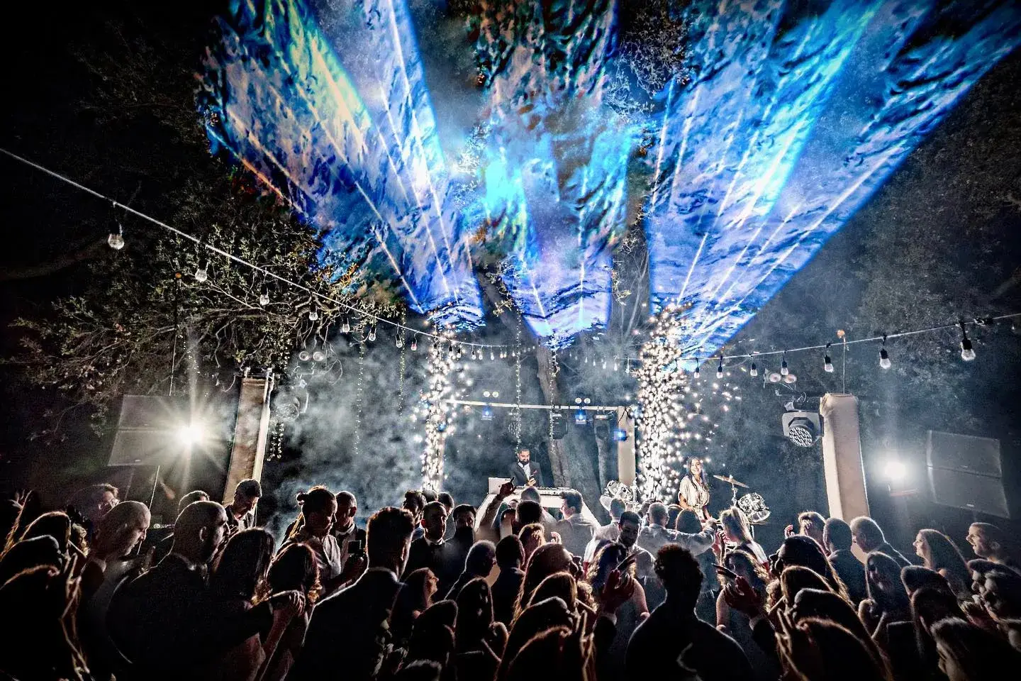 Blue beam lighting and haze over a packed outdoor dance floor, with cold spark fountains at the stage