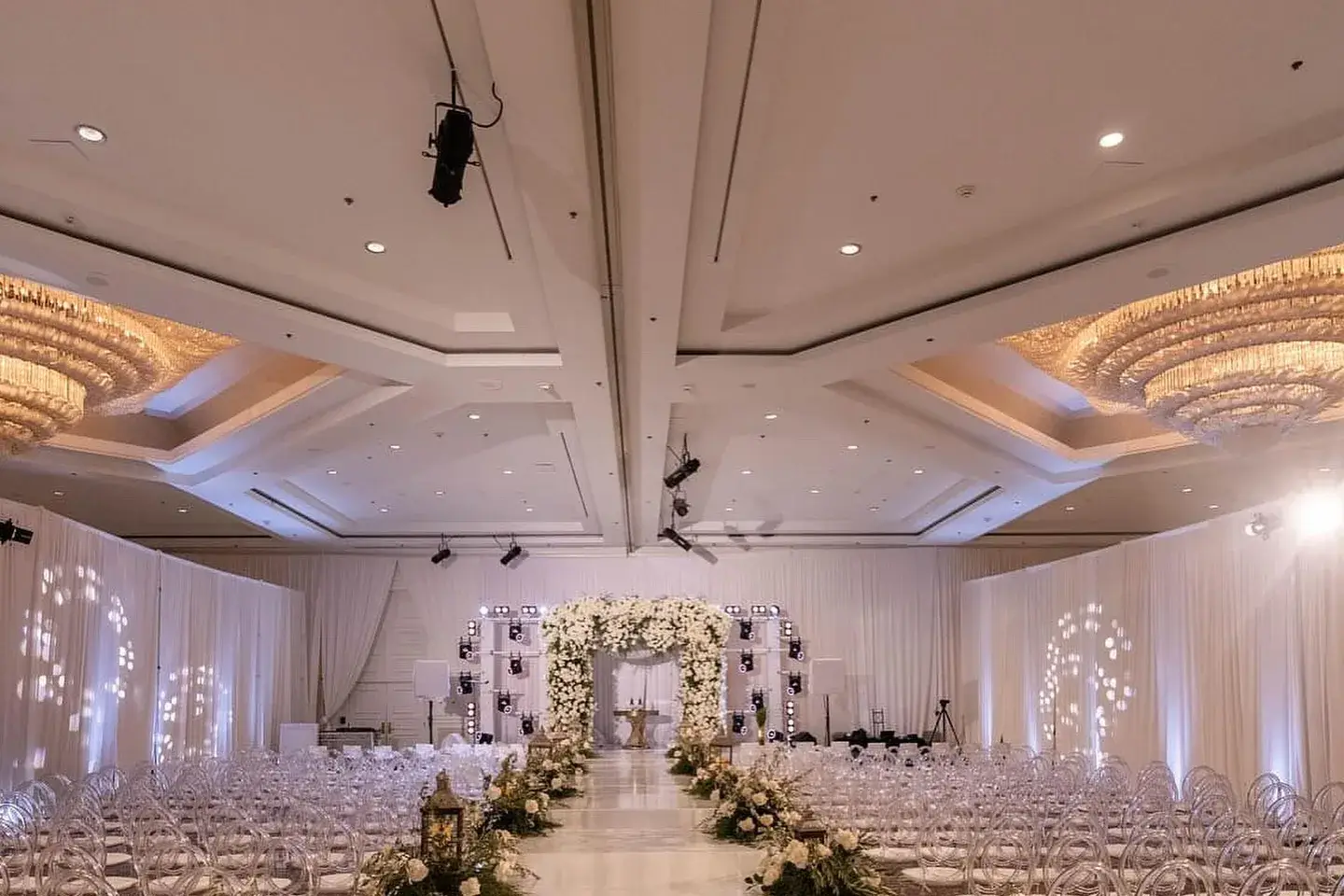 All-white ballroom wedding ceremony with a floral aisle, chandeliers, and soft uplighting
