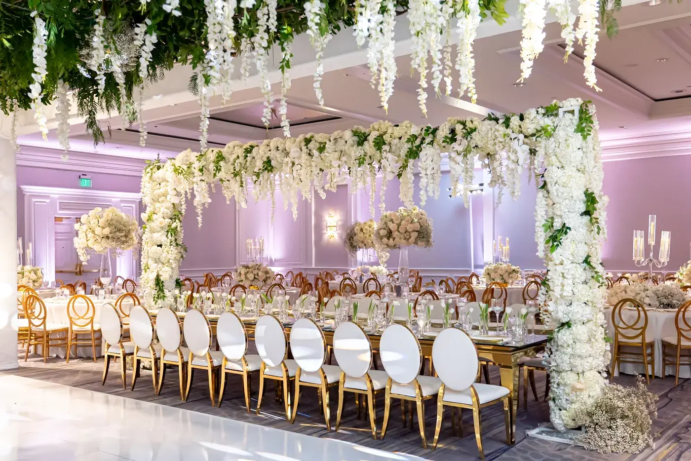 Long gold reception tables under a floral canopy with lavender uplighting at The Ritz-Carlton, Marina del Rey.
