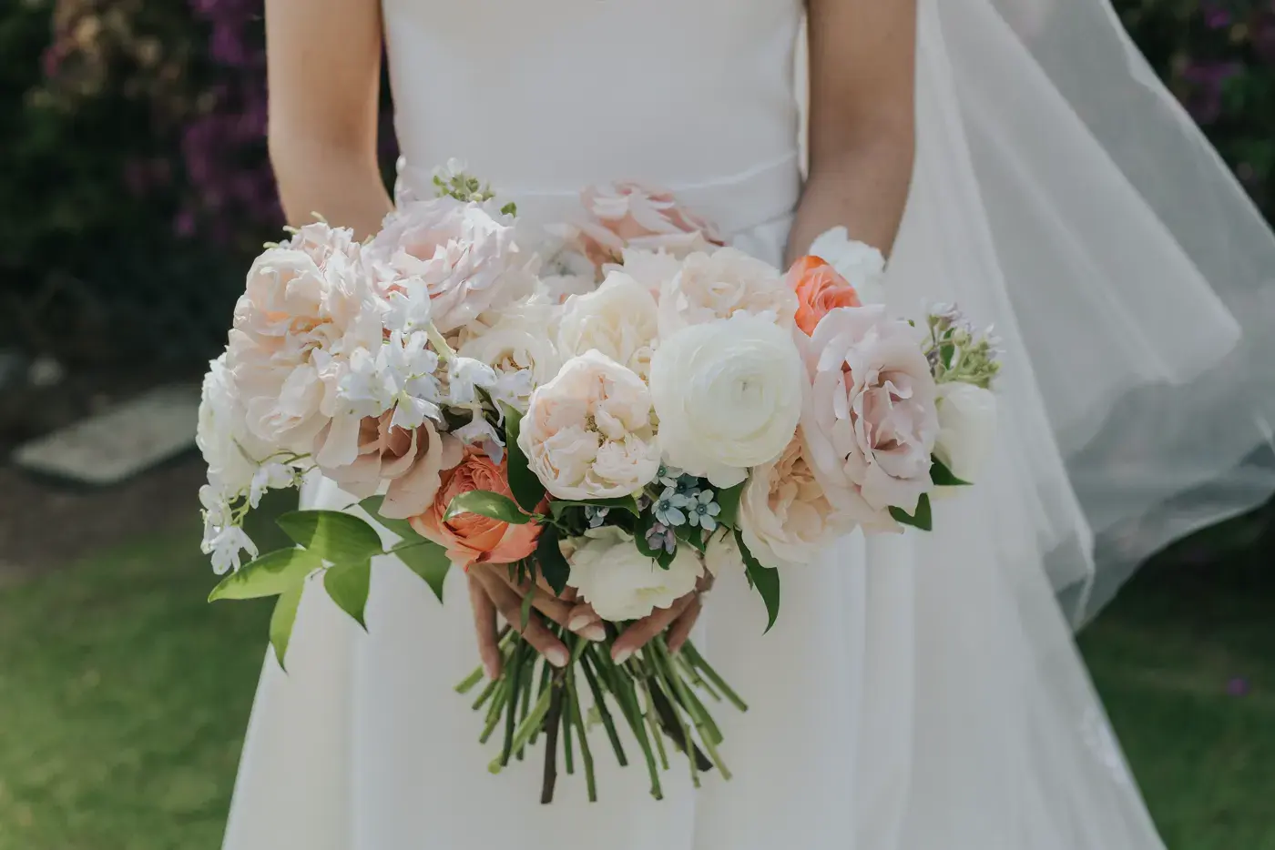 A bridal bouquet of blush and peach roses held against a white gown on the lawn at the Bel-Air Bay Club.