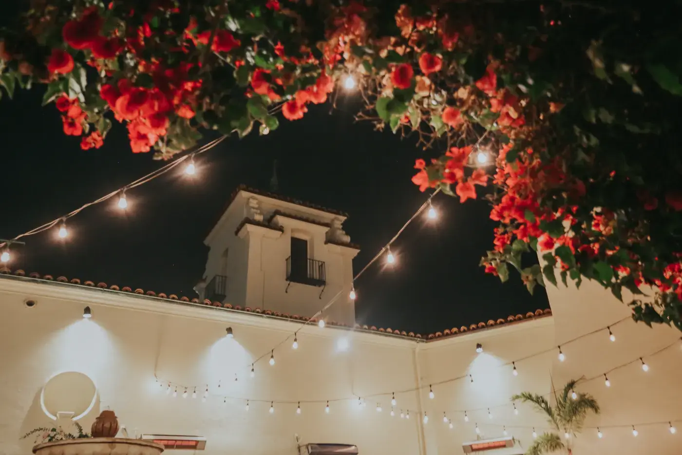 The Bel-Air Bay Club courtyard at night, with string lights crossing overhead, red bougainvillea and the white clubhouse tower.