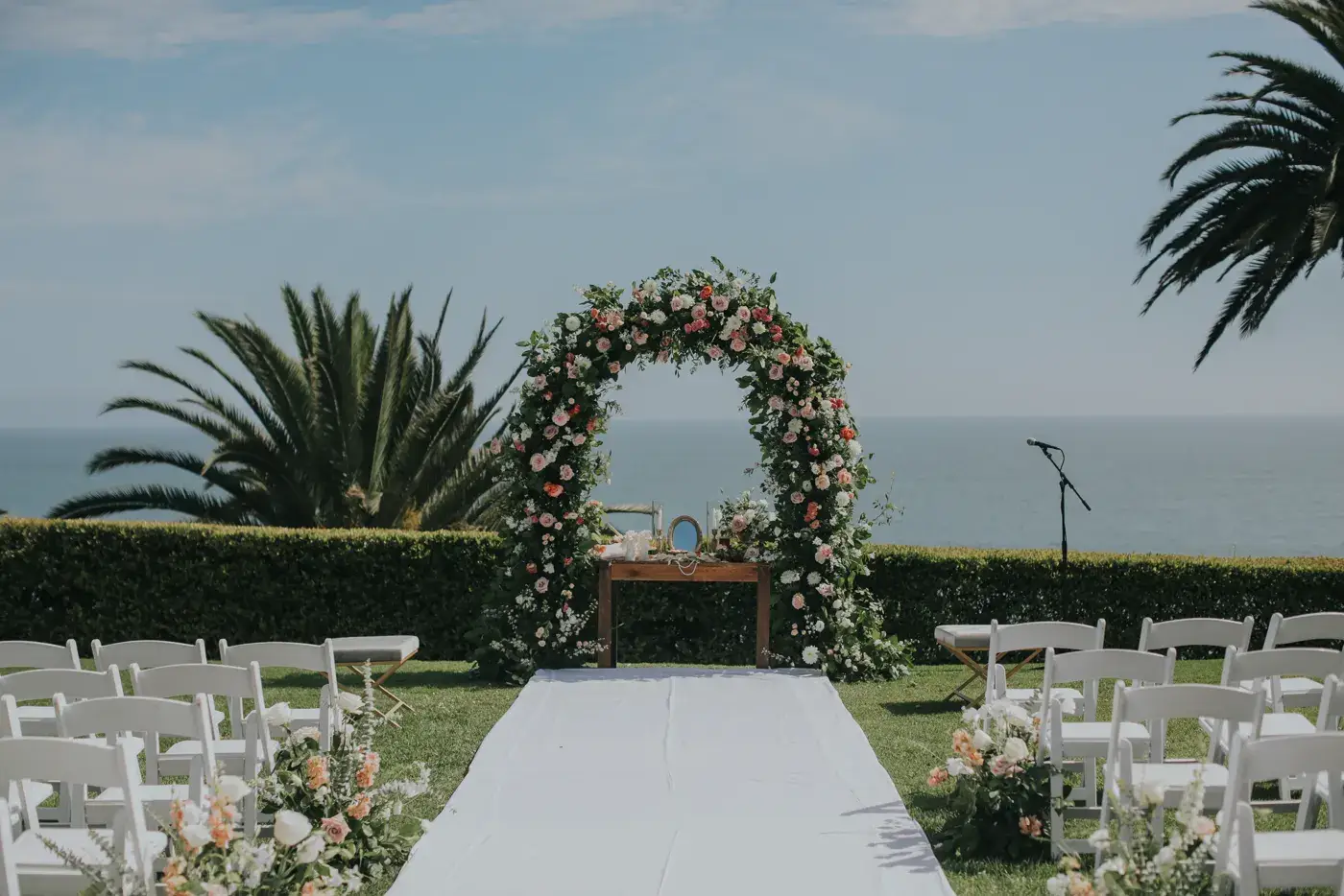 An oceanfront wedding ceremony set at the Bel-Air Bay Club: a white aisle between rows of white chairs, a floral arch and a microphone on a stand, with palm trees and the Pacific beyond.