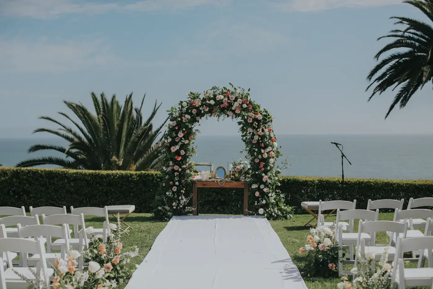 An oceanfront wedding ceremony set at the Bel-Air Bay Club: a white aisle between rows of white chairs, a floral arch and a microphone on a stand, with palm trees and the Pacific beyond.