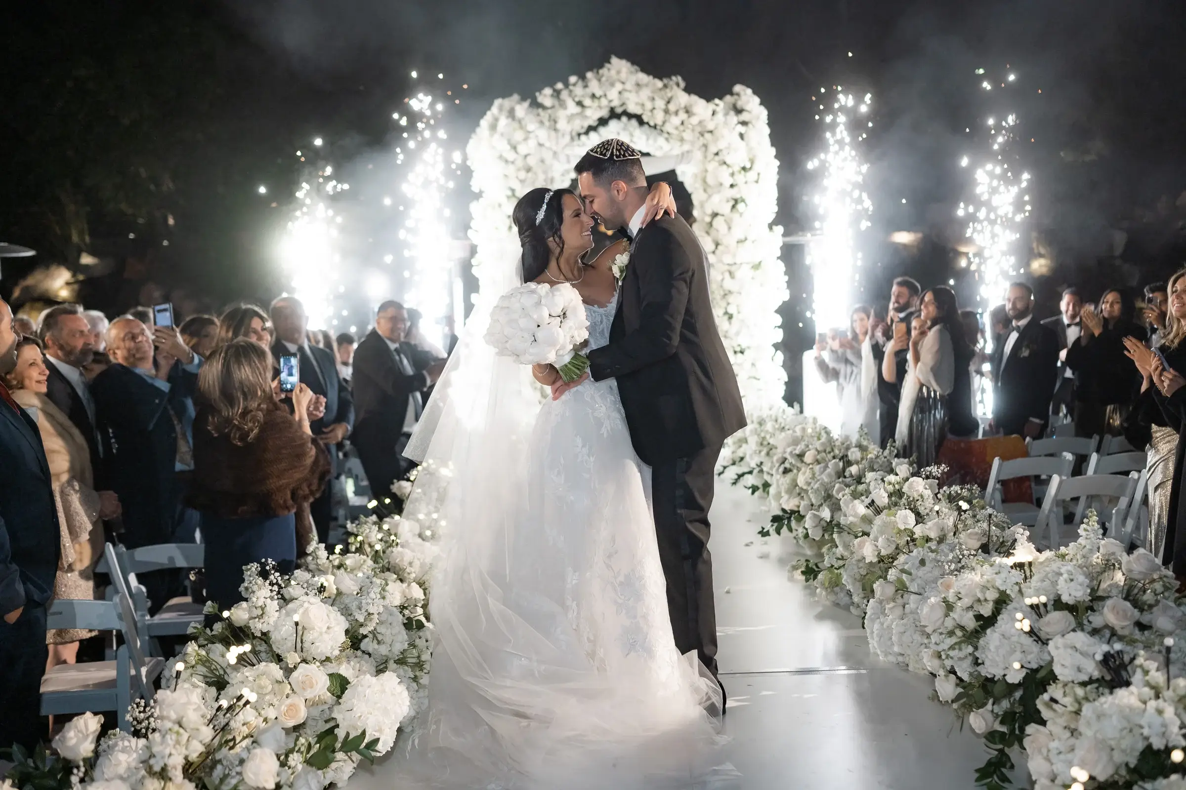 Cold spark fountains lining the aisle as a couple kisses under a white floral arch at the Four Seasons Hotel Westlake Village