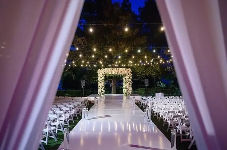 Outdoor evening wedding ceremony with overhead string lights above a white aisle, a white floral chuppah and rows of white chairs